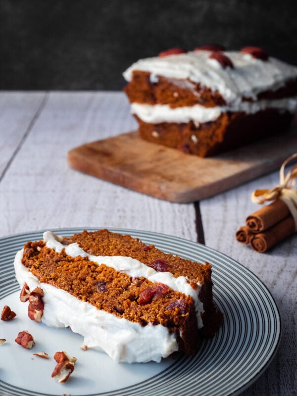 une part de carrot cake dans une assiette à dessert sur une table en bois avec des bâtons de cannelle et le carrot cake sur une planche a découper en arrière plan